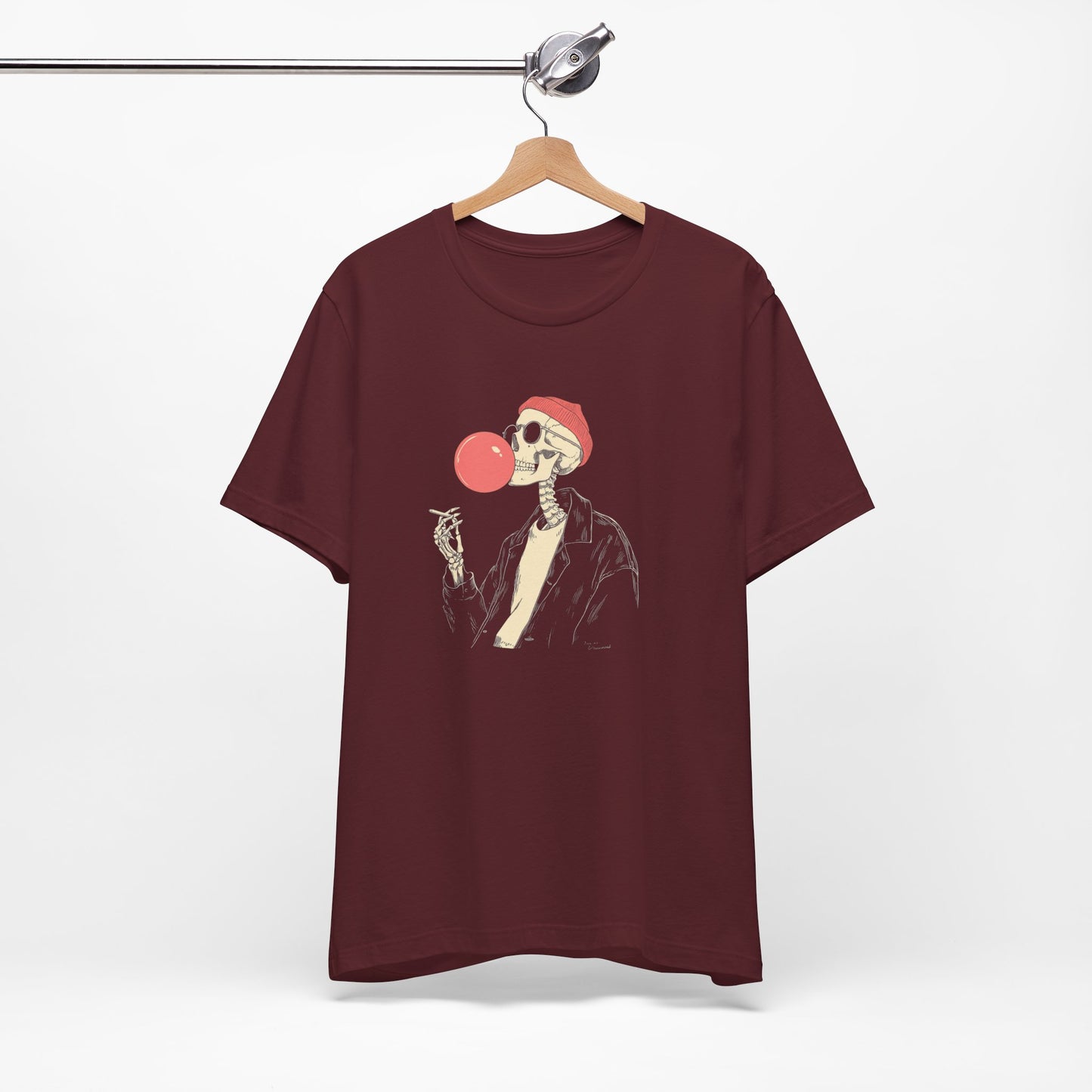 Maroon t-shirt with a skeleton design blowing a bubble gum bubble, hanging on a wooden hanger against a white background.