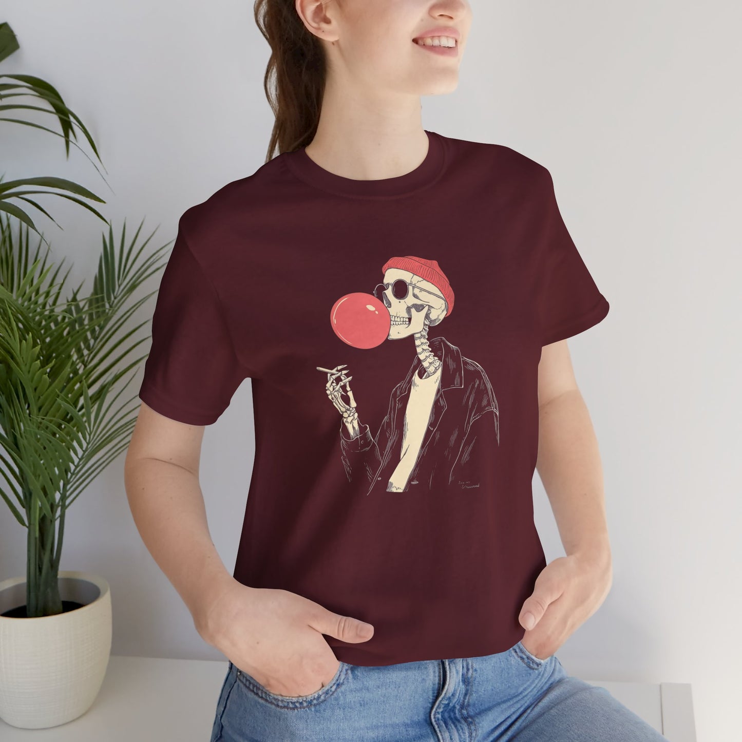 Person wearing a maroon t-shirt with a skeleton design blowing a red bubble gum bubble.