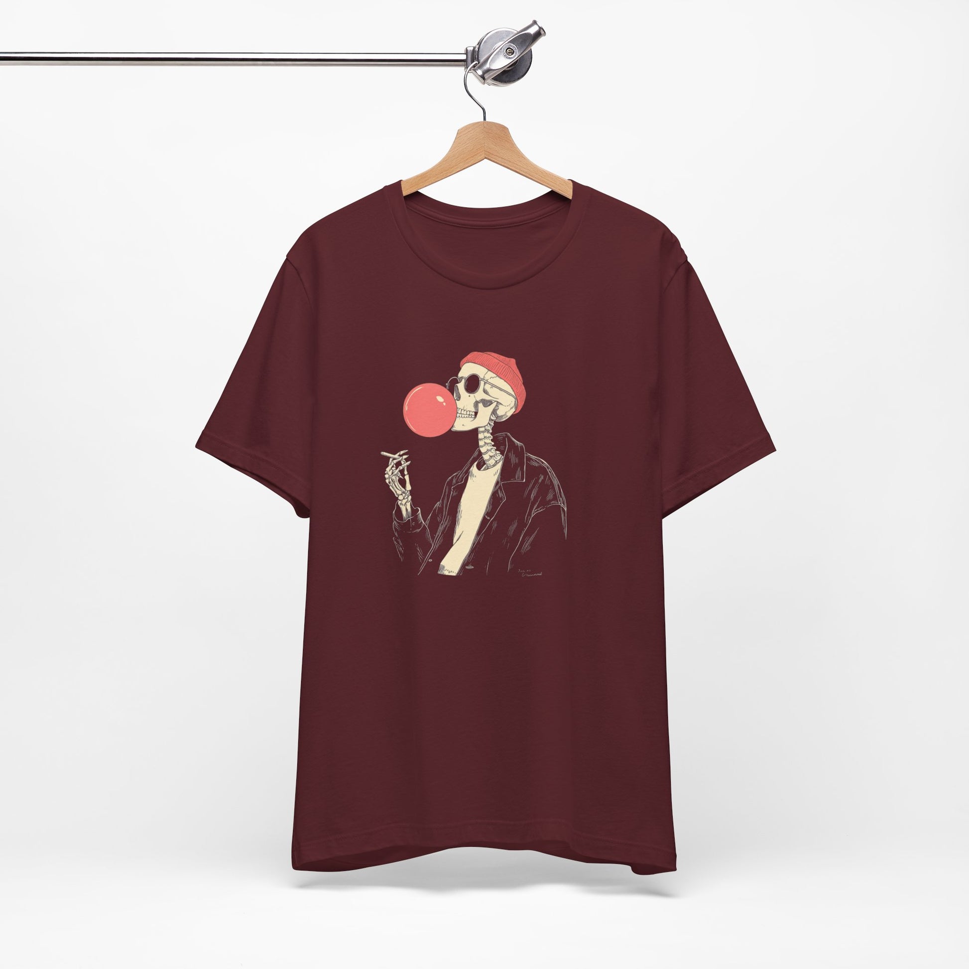 Maroon t-shirt with a skeleton design blowing a bubble gum bubble, hanging on a wooden hanger against a white background.
